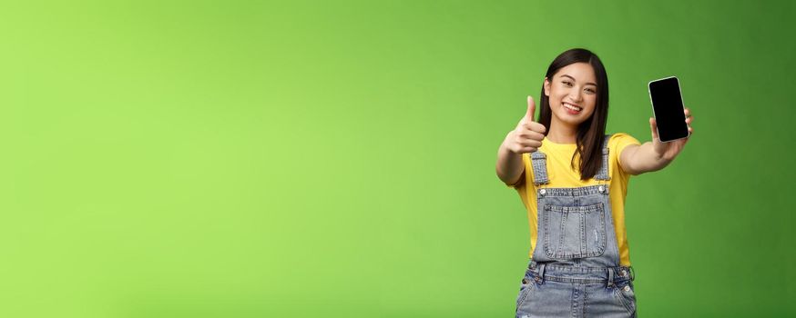 Cheerful satisfied cute asian brunette girl pleased awesome smartphone game, show telephone display, give thumb-up like sign, smiling broadly, introduce awesome online application, green background.