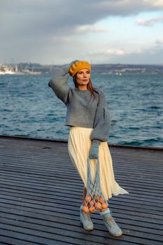 Outdoors fashion portrait of a beautiful middle aged woman walking on the beach. Marine background. Dressed in a stylish warm blue sweater, yellow skirt and beret