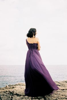 Woman in a long dress fluttering in the wind stands on a rock above the sea. Back view. High quality photo