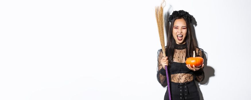 Image of beautiful asian woman dressed-up as a witch for halloween party, holding broom and pumpkin, standing over white background.
