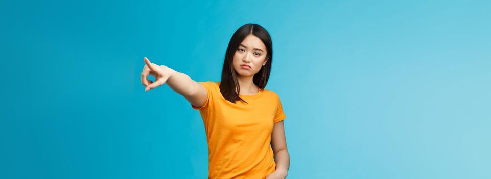 Go away from here. Serious-looking arrogant displeased bossy asian girl showing way-out, pointing sideways index finger look camera demand leave, stand blue background ignorant. Copy space