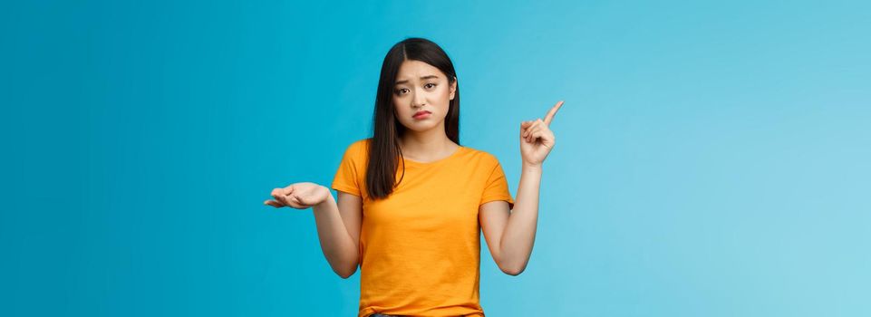 Troubled cute upset asian girl have problem asking your help, pointing upper left corner shrugging unsure, look frustrated and sad, questioned answer, cannot deal problem, stand blue background.
