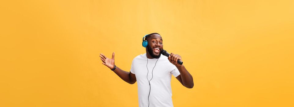 Portrait of cheerful positive chic. handsome african man holding microphone and having headphones on head listening music singing song enjoying weekend vacation isolated on yellow background.