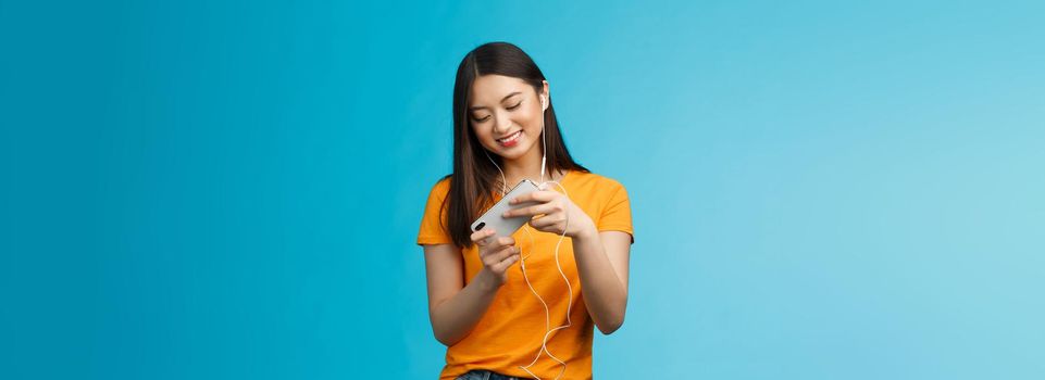 Cute entertained good-looking urban female student spend break playing racing game, hold smartphone horizontal tilt phone move car, smiling broadly standing enthusiastic blue background.