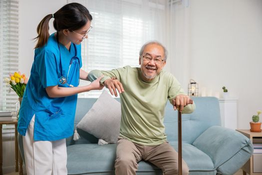 International Day for the Elderly. Young carer supporting helping senior man stand up with walking stick from sofa, Nurse wearing blue uniform help helping his patient senior old man with walking cane