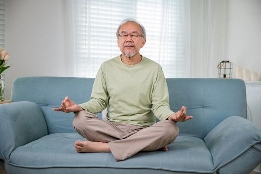 Asian old man practise yoga and meditation in lotus position and closed eyes, lifestyle senior man lotus pose doing yoga for mental balance breathing air relaxing on sofa at home, Healthy life habits