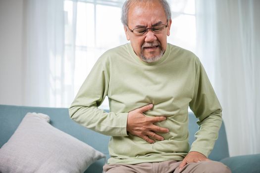 Elderly Asian old man sitting on sofa having stomachache, old age suffering from stomach ache holding his stomach in living room, people health problem, food poisoning