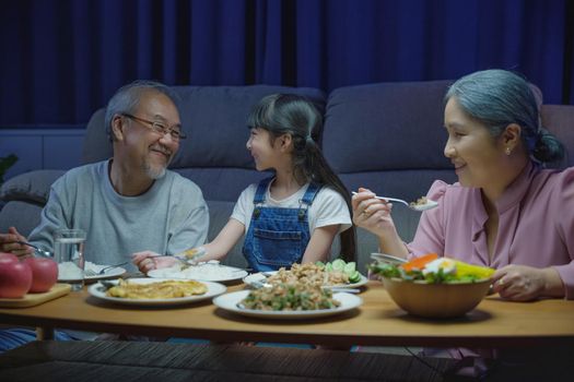 Happy Asian family grandparent and granddaughter dining on table and having fun during at house evening time, Happiness senior parent and child eating dinner food together in living room indoors