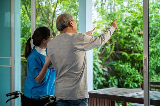 Young Asian nurse is supporting elderly man sitting in wheelchair walking to looking out window on garden and breathe fresh air, pension retirement center, senior man at home, healthcare concept