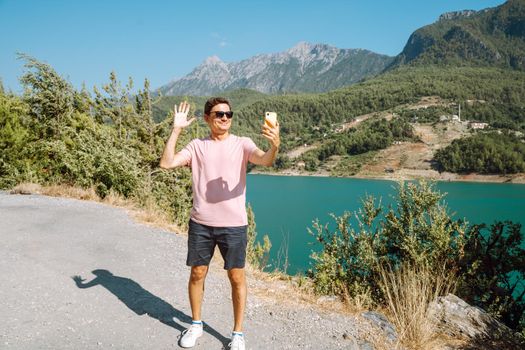 Man taking selfie on mobile phone of mountains lake background. Traveler male having video chat on cellphone on the blue lake outdoors travel adventure vacation.