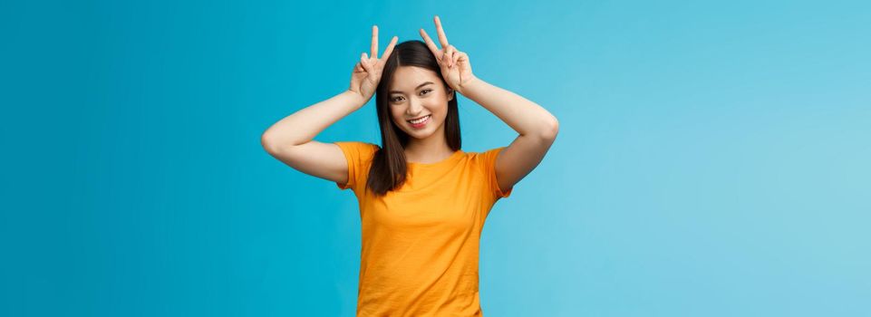 Cute tender asian girlfriend act silly lovely smile, tilt head show playfully ears hold peace victory signs on head, grinning toothy, express joy happiness and delight, stand blue background.