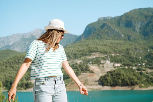 Smiling woman in hat and sunglasses with wild hair standing near mountains lake on background. Positive young woman traveling on blue lake outdoors travel adventure vacation.