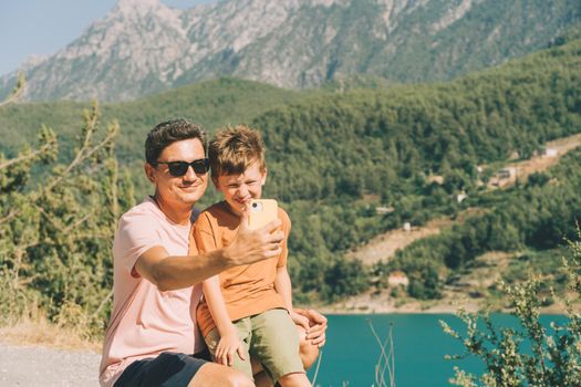 Young dad and his son standing on Mountain View and taking selfie on mobile phone. Child kid boy having fun with father and hiking hiking near mountains lake on background.