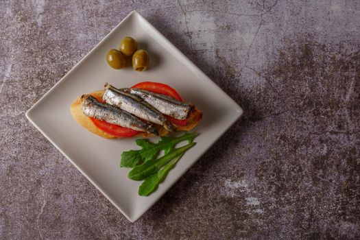 tapa of sardines on a slice of bread with tomato and olives on a white plate with a typical spanish white background