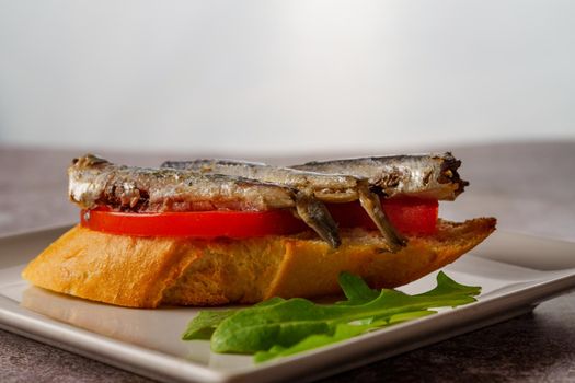 tapa of sardines on a slice of bread with tomato and olives on a white plate with a typical spanish white background