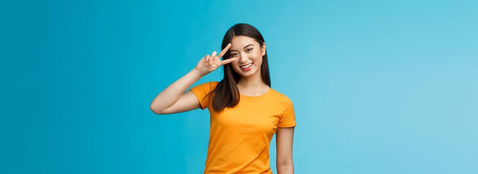 Friendly cheerful asian girlfriend look joy happiness, show peace victory sign, smiling toothy positive, enjoy lucky perfect summer sunny day, stand blue background carefree wear yellow t-shirt.