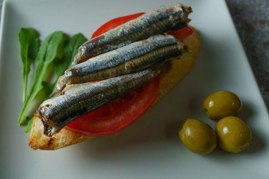 tapa of sardines on a slice of bread with tomato and olives on a white plate with a typical spanish white background