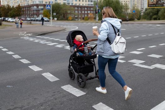 a young mother with a stroller crosses the road at a pedestrian crossing with a cup of coffee in her hands.