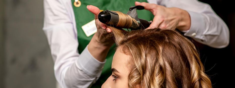 The female hairdresser is curling hair for a brown-haired young caucasian woman in a beauty salon