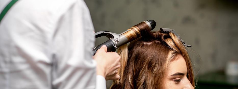 The female hairdresser is curling hair for a brown-haired young caucasian woman in a beauty salon