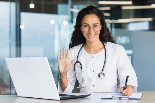 Portrait of a hispanic woman doctor veterinarian. He sits in the office, conducts an online consultation with a laptop, looks and waves at the camera. Records the diagnosis, treatment of the animal.