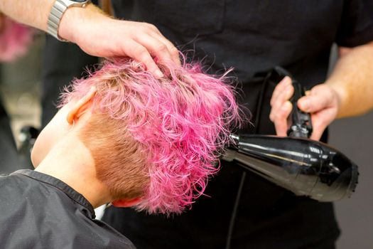 Drying short pink bob hairstyle of a young caucasian woman with a black hair dryer with the brush by hands of a male hairdresser in a hair salon, close up