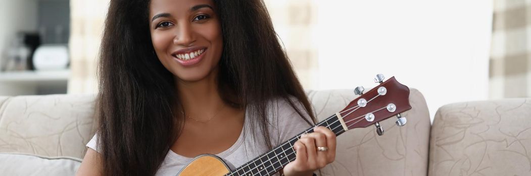Portrait of happy afro american woman playing on ukulele in living room at home, learning new song on musical instrument. Music, hobby, creativity concept