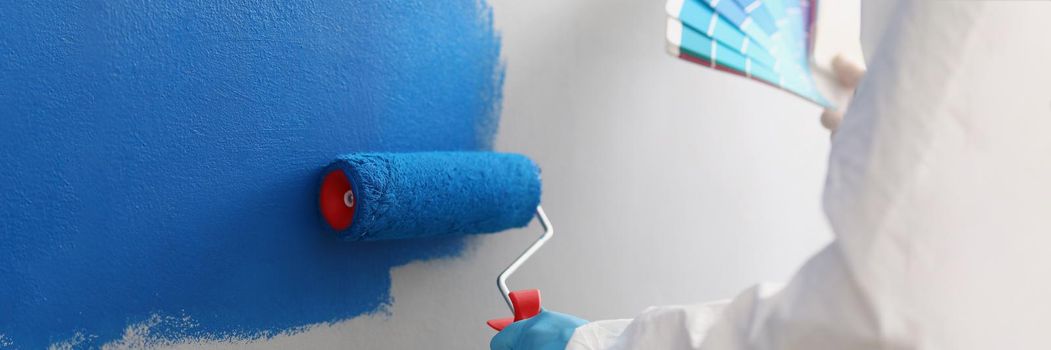 Close-up of painter with brush tool covered in blue colour painting wall, worker on site. Renovation, interior design, construction site, project concept