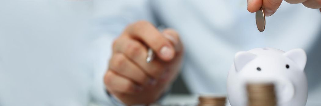 Close-up of person accountant put coin in white piggy bank, stack of coins on workplace. Prepare annual report, count every cent. Economy, savings concept