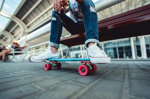 Girl sits on a bench in white sneakers and jeans, Close up of girls feet on a colourfull skateboard.