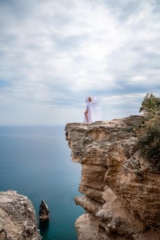 Blonde with long hair on a sunny seashore in a white flowing dress, rear view, silk fabric waving in the wind. Against the backdrop of the blue sky and mountains on the seashore