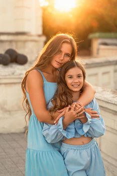 Portrait of mother and daughter in blue dresses with flowing long hair against the backdrop of sunset. The woman hugs and presses the girl to her. They are looking at the camera