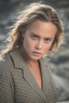 Debonair blonde girl sitting at sandy beach in autumn sunset. Outdoor portrait of pretty curly fashionable girl