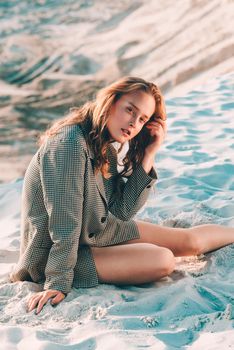 Debonair blonde girl sitting at sandy beach in autumn sunset. Outdoor portrait of pretty curly fashionable girl