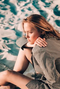 Debonair blonde girl sitting at sandy beach in autumn sunset. Outdoor portrait of pretty curly fashionable girl