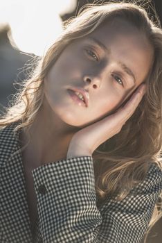 Debonair blonde girl sitting at sandy beach in autumn sunset. Outdoor portrait of pretty curly fashionable girl