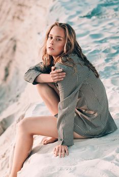 Debonair blonde girl sitting at sandy beach in autumn sunset. Outdoor portrait of pretty curly fashionable girl