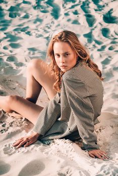 Debonair blonde girl sitting at sandy beach in autumn sunset. Outdoor portrait of pretty curly fashionable girl