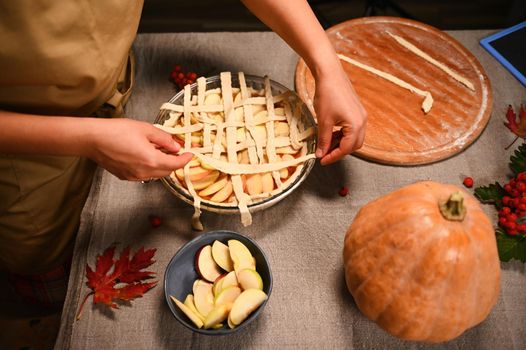 Top view of a housewife in beige chef's apron, decorating the classic American homemade festive pie with a crunchy pastry lattice. Thanksgiving Day. Making tasty pumpkin apple pie with flaky crust.