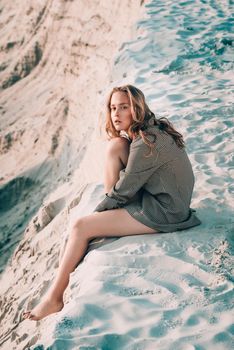 Debonair blonde girl sitting at sandy beach in autumn sunset. Outdoor portrait of pretty curly fashionable girl