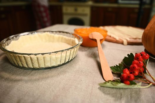 Still life with a rolled out pastry dough in a baking dish, a bowl with pumpkin puree, a branch with viburnum berries on a table with linen tablecloth. Thanksgiving Day and Halloween concept