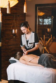 Young charming girl on a panchakarma procedure laying on a massage table. beautiful woman spending time at modern spa cabinet relaxing. Soft yellow light.