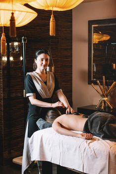 Specialist making hot stone massage to a client. Young charming girl on a panchakarma procedure laying on a massage table. beautiful woman spending time at modern spa cabinet relaxing. Soft yellow light.