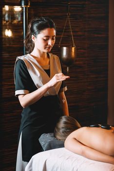 Specialist making biolocation with crystal pendulum. Young charming girl on a panchakarma procedure laying on a massage table. beautiful woman spending time at modern spa cabinet relaxing. Soft yellow light.