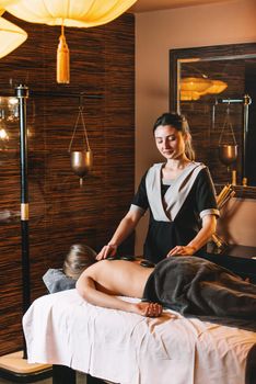 Specialist making hot stone massage to a client. Young charming girl on a panchakarma procedure laying on a massage table. beautiful woman spending time at modern spa cabinet relaxing. Soft yellow light.