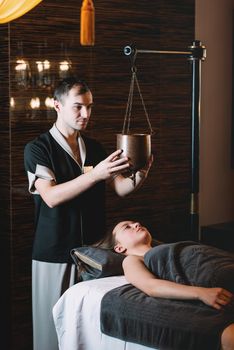 Specialist making shirodhara massage form of ayurveda therapy. Young charming girl on a panchakarma procedure laying on a massage table. beautiful woman spending time at modern spa cabinet relaxing. Soft yellow light.