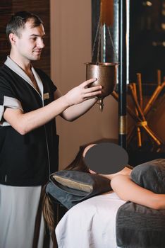 Specialist making shirodhara massage form of ayurveda therapy. Young charming girl on a panchakarma procedure laying on a massage table. beautiful woman spending time at modern spa cabinet relaxing. Soft yellow light.