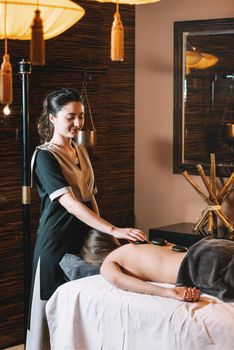 Specialist making hot stone massage to a client. Young charming girl on a panchakarma procedure laying on a massage table. beautiful woman spending time at modern spa cabinet relaxing. Soft yellow light.