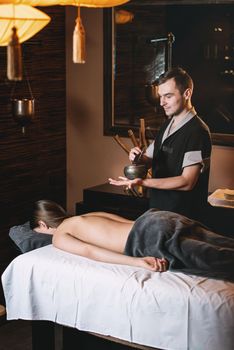 Specialist holding in hands Brass Hammer Singing Bowl. Young charming girl on a panchakarma procedure laying on a massage table. beautiful woman spending time at modern spa cabinet relaxing. Soft yellow light.