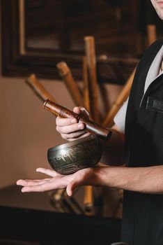 Specialist holding in hands Brass Hammer Singing Bowl. Young charming girl on a panchakarma procedure laying on a massage table. beautiful woman spending time at modern spa cabinet relaxing. Soft yellow light.